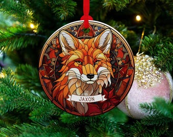 May include: A round ceramic ornament featuring a stained-glass style illustration of a fox in shades of orange, red, and brown. The design includes a white banner at the bottom with the name JAXON printed in dark letters, suspended by a red ribbon.
