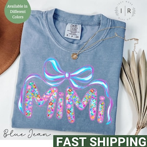 May include: A blue jean t-shirt with the word "Mimi" in pink letters outlined with colorful flowers and a pink bow with blue and white ribbon. The shirt has a tag that says "Comfort Colors" and "S-P-CH".