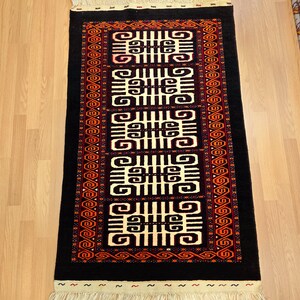 Puede incluir: Una alfombra rectangular negra con un patrón central de formas geométricas color crema. La alfombra tiene un borde rojo y naranja con un diseño repetido y flecos color crema en ambos extremos. Está sobre un suelo de madera marrón claro.