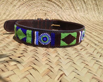 May include: A brown leather dog collar with a beaded design. The collar features blue, green, white, and brown beads in geometric patterns. The collar has a gold-colored buckle and is set against a woven background.
