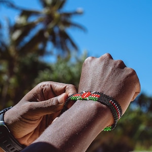 Könnte beinhalten: Ein Armband aus Perlen mit roten, weißen, grünen und schwarzen Perlen. Das Armband wird am Handgelenk befestigt. Der Hintergrund umfasst einen hellblauen Himmel und Palmen.