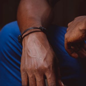 May include: A dark brown leather cord bracelet featuring two thin strands and a black metal clasp. The bracelet is worn around the wrist, showcasing a simple design with a smooth texture and a secure fastening mechanism that connects the two leather cords.