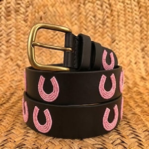 May include: A black leather belt featuring a series of light pink beaded horseshoe motifs along the strap. The belt is secured with a gold-toned metal buckle and includes two matching leather loops to hold the end of the strap in place.