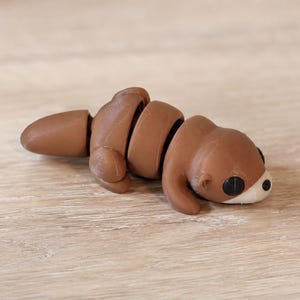 May include: A small, articulated brown otter figurine with a smooth, matte finish. The creature features a segmented body, a rounded tail, small limbs, and a light-colored snout with black eyes and a black nose, designed with visible joints.