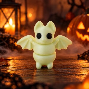 May include: A small, pale yellow, glow-in-the-dark bat figurine with large, round, solid black eyes. The bat has pointed ears, outstretched wings, and a rounded body, standing upright on a surface with a smooth, matte texture.