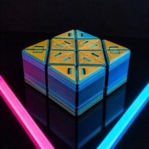 May include: A multi-layered puzzle cube featuring a geometric design with gold-colored top surfaces and blue-toned sides. The cube displays a textured finish on its top panels, which are divided into triangular and square segments, creating a complex pattern.