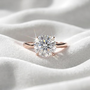 May include: A solitaire ring featuring a large, round, brilliant-cut clear gemstone set in a four-prong rose gold band. The center stone exhibits significant light refraction and sparkle, creating a starburst effect against the metal setting.