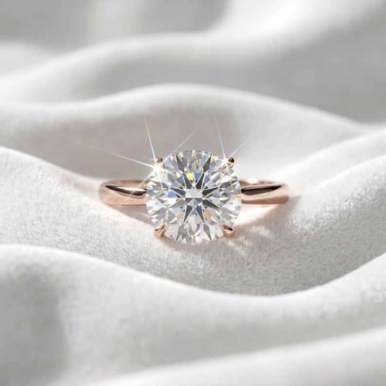 May include: A solitaire ring featuring a large, round, brilliant-cut clear gemstone set in a four-prong rose gold band. The center stone exhibits significant light refraction and sparkle, creating a starburst effect against the metal setting.