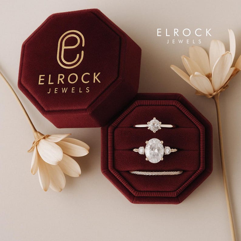 May include: A burgundy velvet ring box with a gold logo that says "Elrock Jewels" and a gold "E" inside a circle. The box is open and contains three rings, one with a large oval-shaped diamond and two smaller diamond rings.