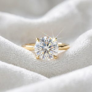 May include: A gold ring featuring a large, round, brilliant-cut clear gemstone held by four prongs. The stone exhibits a bright, star-like light reflection, and the band is a smooth, polished yellow gold metal that rests on a soft white fabric.