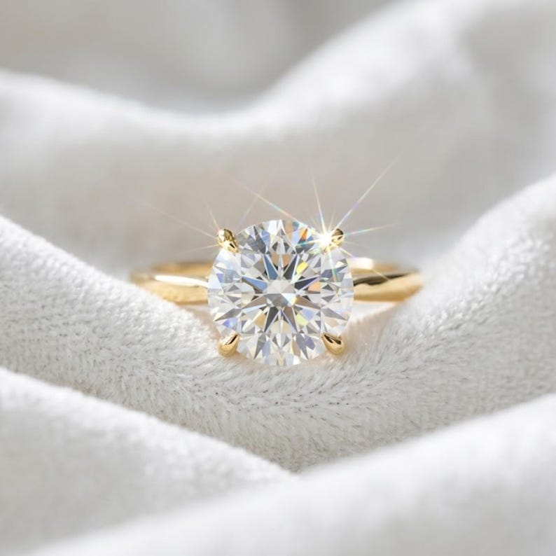 May include: A gold ring featuring a large, round, brilliant-cut clear gemstone held by four prongs. The stone exhibits a bright, star-like light reflection, and the band is a smooth, polished yellow gold metal that rests on a soft white fabric.