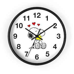 May include: A round wall clock with a black frame and a white face featuring black numbers from 1 to 12. The center displays a graphic of a white dog hugging a small yellow bird, surrounded by three small red hearts above their heads.