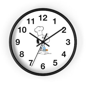 May include: A round wall clock with a black frame and a white face featuring black numbers from 1 to 12. In the center, a cartoon dog wearing a white chef hat and a light blue apron holds an orange wooden spoon. The clock has black hour, minute, and second hands.