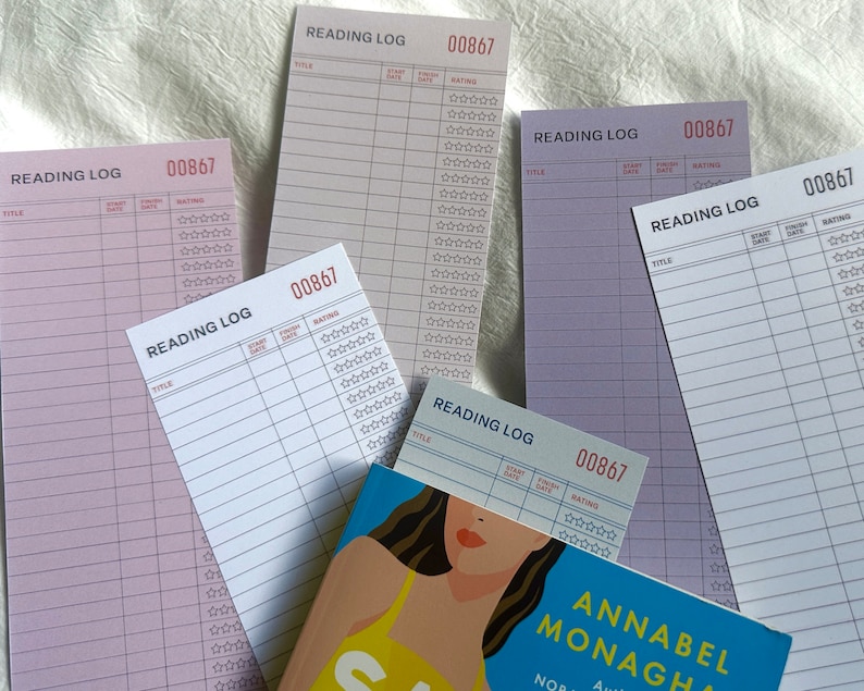 May include: Five reading log notepads with a pink and white striped design. Each notepad has a title, start date, finish date, and rating section. The notepads are labeled "Reading Log" and have a number "00867" printed on them. A book with a colorful cover is in the foreground.