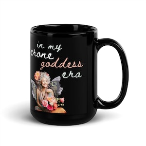 May include: Black ceramic mug with a white design that says "in my crone goddess era". The design also features a woman with gray hair and wings, holding flowers.