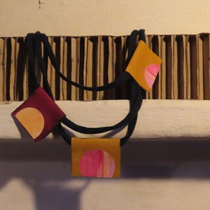 May include: A unique necklace featuring a black cord and three square pendants. The pendants are in shades of burgundy and mustard yellow, each adorned with a painted circular design in complementary colors. The necklace is a statement piece.
