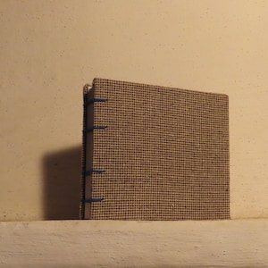 Op de afbeelding: Een vierkant, handgemaakt notitieboek met een textuur, bruine stoffen kaft. De rug is gebonden met blauw garen. Het notitieboek ligt op een witte richel tegen een beige muur.