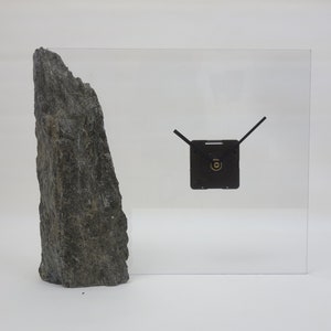 Puede incluir: Una roca gris se encuentra junto a una esfera de reloj de acrílico transparente con manecillas negras. La esfera del reloj es cuadrada y tiene un pequeño círculo dorado en el centro.