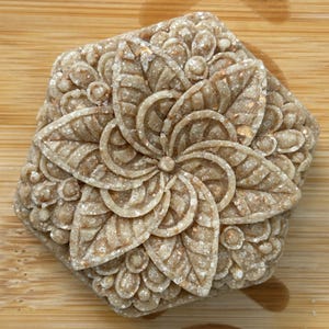 May include: A square, light brown soap bar with a detailed floral design. The soap features a central flower with large, textured petals and is surrounded by smaller, circular embellishments. The bar is set on a wooden surface.