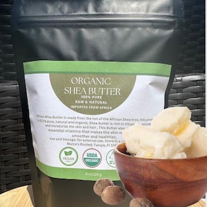 May include: A white bag with a green label that reads "ORGANIC SHEA BUTTER 100% PURE RAW & NATURAL IMPORTED FROM AFRICA". The label also includes a USDA Organic seal and a Vegan seal. The bag is sitting on a brown wicker surface. There is a wooden bowl with shea butter in it.