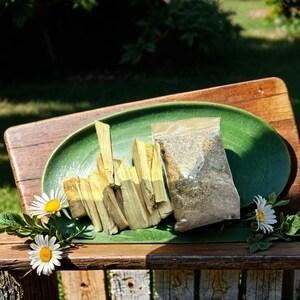 Peut inclure: Une assiette en céramique verte avec un tas de bâtonnets brun clair et un sac en plastique transparent rempli de poudre brune. L'assiette est posée sur une surface en bois avec quelques marguerites blanches.