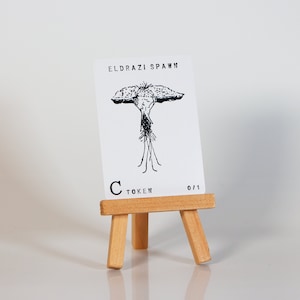 May include: A white card with black text and a black and white illustration of a creature with a large head and many legs. The card reads "Eldrazi Spawn" and "C Token 0/1". The card is on a small wooden easel.