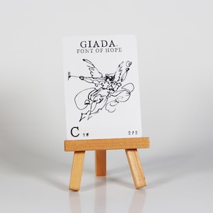 May include: A white card with black ink depicting an angel with wings outstretched, holding a small object in its right hand. The card reads "GIADA. FONT OF HOPE" and "C 1W 2/2". The card is displayed on a small wooden easel.