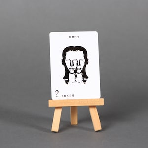 May include: A white card with black text and a black and white illustration of a face. The card is on a small wooden easel. The text on the card reads "COPY ? TOKEN".