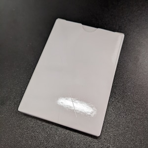 Könnte beinhalten: Ein weißer, rechteckiger Kartenhalter mit leicht abgerundeter Oberkante. Der Halter ist aus glänzendem Material gefertigt und hat eine leichte Falte in der Mitte.