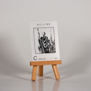 May include: A black and white illustration of a skeleton holding a staff, printed on a white card with the text "SKELETON C TOKEN 4/11". The card is displayed on a small wooden easel.