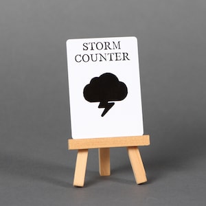 May include: A white card with the words "STORM COUNTER" and a black cloud with a lightning bolt icon, displayed on a small wooden easel. The background is a solid gray.