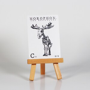 May include: A black and white illustration of a moose skeleton on a white card with the text "MOROPHON. THE BOUNDLESS" and "C 7 6/6". The card is displayed on a small wooden easel.