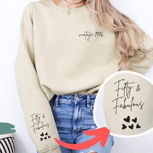 May include: A sand-coloured crewneck sweatshirt featuring black text on the upper left chest that reads vintage 1976. The right sleeve displays the black text Fifty & Fabulous above a cluster of four small black heart icons in a script font.