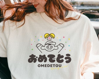 Könnte beinhalten: Ein weißes T-Shirt mit einer Cartoon-Illustration einer Person, die einen Hut trägt und ein Geschenk hält. Der Text "omedetou" ist unter der Illustration gedruckt.
