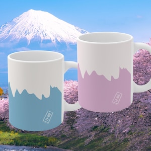 May include: Two white ceramic mugs with a mountain-like design in blue and pink. Each mug has a white handle and a small Japanese character stamp. The background features a mountain and cherry blossoms.