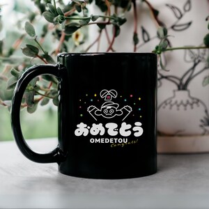 Peut inclure: Mug en céramique noire avec une illustration blanche d'un personnage de dessin animé et le mot japonais "Omedetou" et le mot anglais "Congrats!". La tasse a une anse incurvée.