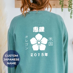 May include: A teal sweatshirt with a white floral design and Japanese text. The back of the sweatshirt features the words "Oregon" and "2015" in white. A circular graphic at the bottom says "Custom Japanese Name".