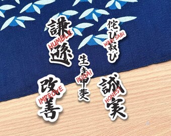 May include: A collection of Japanese-style stickers with black and red text on a white background. The stickers are placed on a blue patterned fabric and a wooden surface. The text includes words like "Humble", "Wabi Sabi", "Ikigai", "Improve", and "Honest".