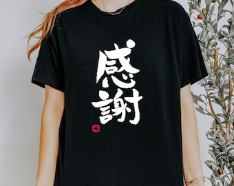 May include: Black t-shirt featuring the Japanese characters for "Thank You" in white, arranged vertically. A small red square with a symbol is at the bottom. The shirt has a crew neck.