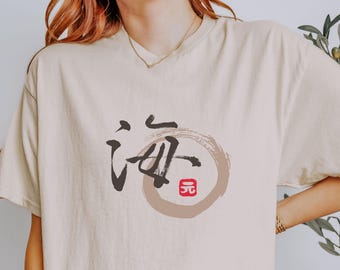 May include: A beige t-shirt with a Japanese calligraphy design. The design features a dark brown character inside a light brown circle, with a small red square at the bottom. The shirt is a neutral color.