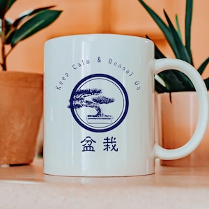 Könnte beinhalten: Eine weiße Keramiktasse mit einem blauen Design, auf dem "Keep Calm & Bonsai On" steht, mit einer Bonsai-Baumillustration und den japanischen Schriftzeichen für Bonsai.