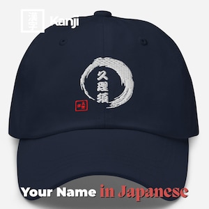 以下が含まれることがあります： 紺色のベースボールキャップ。白い刺繍で円の中に日本の漢字が描かれています。「Your Name in Japanese」の文字が下部にプリントされています。キャップはカーブしたつばと、布製のストラップ留めが付いています。