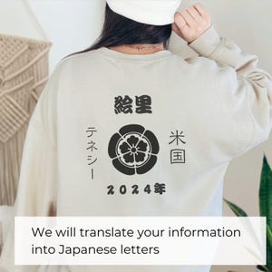 以下が含まれることがあります： 花柄の黒いプリントと黒い文字が書かれたベージュのトレーナー。文字は「絵里」「テネシー」「米国」「2024年」と書いてある。