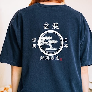 May include: Navy blue t-shirt with a white graphic of a bonsai tree inside a circle, with Japanese text above and to the sides. The shirt has short sleeves and a relaxed fit.