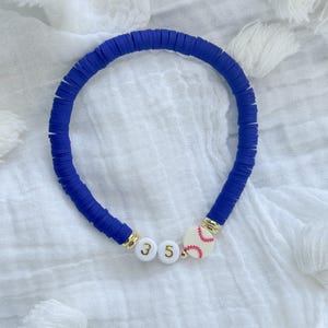 Könnte beinhalten: Ein blaues Armband mit Scheibenperlen, goldenen Akzenten und weißen Buchstabenperlen mit der Aufschrift "35". Ein Baseball-Charm mit roten Nähten ist ebenfalls enthalten. Das Armband wird auf einer weißen, strukturierten Oberfläche präsentiert.