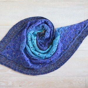 Może przedstawiać: Ręcznie robiona chusta w odcieniach niebieskiego, przechodząca od głębokiego indygo do jasnego turkusu. Chusta posiada skomplikowane wzory warkoczy i koronek, tworząc teksturowany i atrakcyjny wizualnie design. Przedmiot jest prezentowany na jasnej drewnianej powierzchni.