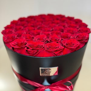 May include: A black round box filled with red roses, tied with a red ribbon. The box has a silver label with the word "Foreverly" on it.