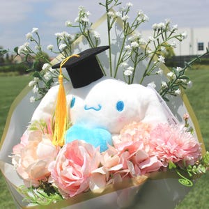 May include: A graduation bouquet featuring a white plush character wearing a black mortarboard with a yellow tassel. The bouquet includes pink and peach-colored flowers, baby's breath, and a blue accent.