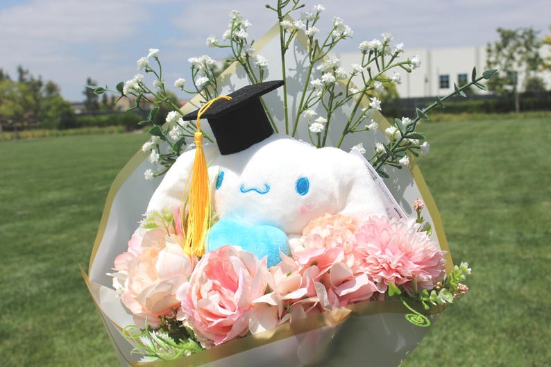 May include: A graduation bouquet featuring a white plush character wearing a black mortarboard with a yellow tassel. The bouquet includes pink and peach-colored flowers, baby's breath, and a blue accent.