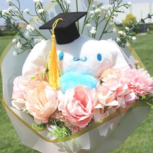 May include: A plush white character wearing a graduation cap, surrounded by pink and peach flowers, and white baby's breath, wrapped in decorative paper. The bouquet has a blue heart-shaped accent and a yellow tassel.